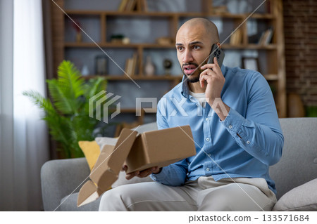 Man on couch holding a damaged cardboard package while making a phone call to customer service, expressing frustration over a faulty online order and problematic delivery Man on couch holding a damaged cardboard package while making a phone call to customer service, expressing frustration over a faulty online order and problematic delivery 133571684