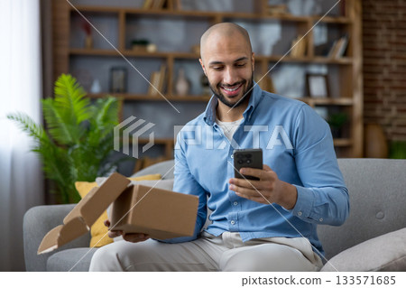 Smiling man unboxing a cardboard package on his couch at home while checking his smartphone after online shopping, relaxed and satisfied with his new purchase and delivery 133571685
