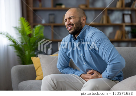Man experiencing intense abdominal pain and discomfort, sitting on a living room couch and pressing hands against his stomach due to digestive health issues or gastritis Man experiencing intense abdominal pain and discomfort, sitting on a living room couch and pressing hands against his stomach due to digestive health issues or gastritis 133571688
