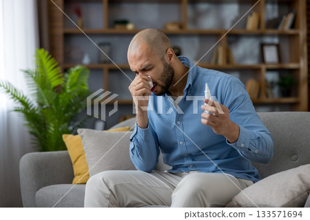 Adult man sitting on a couch at home, blowing his nose with a tissue and holding a nasal spray, experiencing seasonal allergies, flu, or cold symptoms 133571694