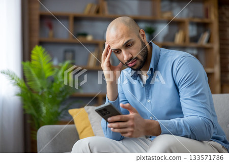 Man with worried expression sitting on sofa, holding smartphone and staring at screen, stressed and anxious after receiving bad news, tense domestic moment of modern life Man with worried expression sitting on sofa, holding smartphone and staring at screen, stressed and anxious after receiving bad news, tense domestic moment of modern life 133571786