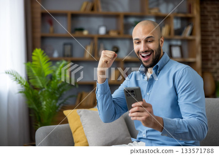 Young man on a couch in a modern living room joyfully checks his smartphone, celebrating a win or good news with a smile and triumphant gesture of excitement Young man on a couch in a modern living room joyfully checks his smartphone, celebrating a win or good news with a smile and triumphant gesture of excitement 133571788