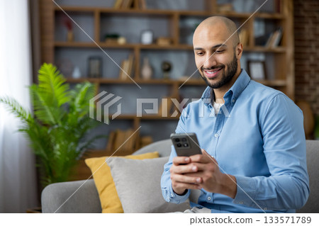 Young man smiling and relaxing on a couch at home, browsing social media on his smartphone while engaging with digital content, representing modern communication and leisure 133571789