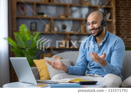 Man wearing an office headset is having a video call on a laptop from his living room sofa, actively communicating and remote working in a modern home office setup Man wearing an office headset is having a video call on a laptop from his living room sofa, actively communicating and remote working in a modern home office setup 133571791