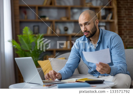 Young man analyzing financial documents and using a laptop while working from home, focusing on budgeting, bill payment, and managing remote work responsibilities Young man analyzing financial documents and using a laptop while working from home, focusing on budgeting, bill payment, and managing remote work responsibilities 133571792
