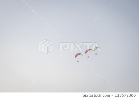 Brightly colored paramotors fly in tight groups across a clear, calm sky during a daytime adventure. 133572300