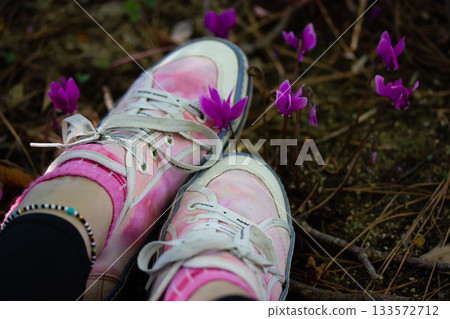 Stylish girl in pink sneakers sitting among purple violet cyclamen hederifolium flowers in the garden. Oneness with nature. Travel discover the World. 133572712