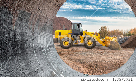 Heavy machinery operates on a construction site, shaping the ground and moving materials while surrounded by hills and clear blue skies during midday 133573731