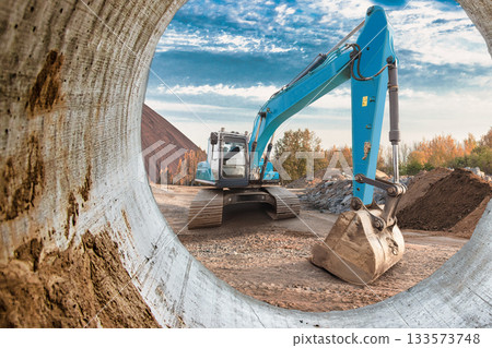 A blue excavator is digging at a construction site surrounded by dirt piles and trees under a bright sky during the day A blue excavator is digging at a construction site surrounded by dirt piles and trees under a bright sky during the day 133573748