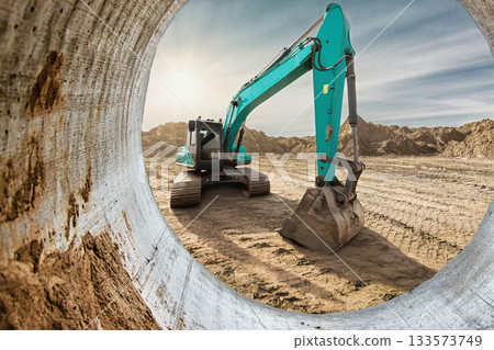 A bright blue excavator operates at a construction site, digging into the earth as the sun sets on the horizon, creating a warm glow around the scene A bright blue excavator operates at a construction site, digging into the earth as the sun sets on the horizon, creating a warm glow around the scene 133573749