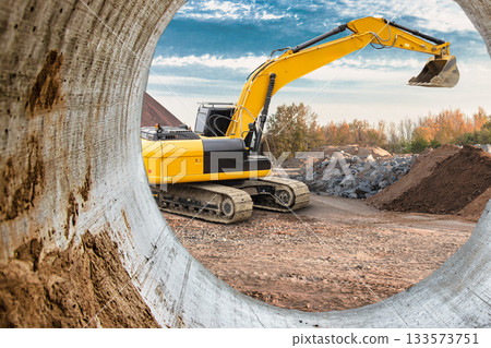 Heavy machinery is actively digging at a construction site, framed by a concrete pipe, with autumn trees in the background under a clear sky Heavy machinery is actively digging at a construction site, framed by a concrete pipe, with autumn trees in the background under a clear sky 133573751