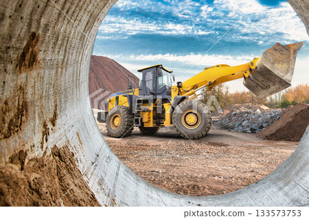 A large yellow loader is actively moving dirt and materials at a construction site. The backdrop includes lush trees and distant hills under a blue sky 133573753