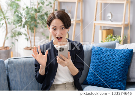 A woman operating a smartphone on the sofa is surprised A woman operating a smartphone on the sofa is surprised 133574646