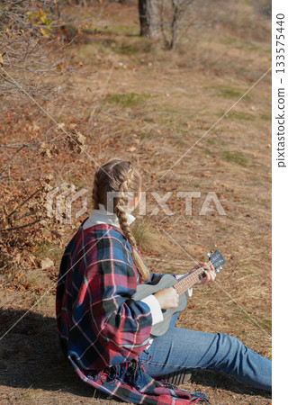 Blonde woman playing guitar ukulele outdoors in autumn forest. Concept of sound therapy, mental health and wellness rituals. Calmness tranquility audio-sensory practices. Aura farming 133575440