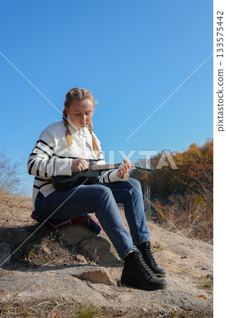 Beautiful blonde girl playing guitar ukulele and singing relaxing enjoying outdoors on sunny autumn day. Hippie woman in boho style training playing musical instrument. Fall leaves 133575442