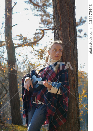 Blonde woman playing guitar ukulele outdoors in autumn forest. Concept of sound therapy, mental health and wellness rituals. Calmness tranquility audio-sensory practices. Aura farming Blonde woman playing guitar ukulele outdoors in autumn forest. Concept of sound therapy, mental health and wellness rituals. Calmness tranquility audio-sensory practices. Aura farming 133575448