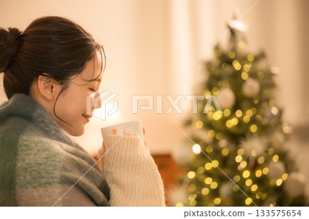 一位年輕女子在夜晚的房間裡喝著熱飲。冬日景象,一位女子與聖誕樹相伴。 一位年輕女子在夜晚的房間裡喝著熱飲。冬日景象,一位女子與聖誕樹相伴。 133575654