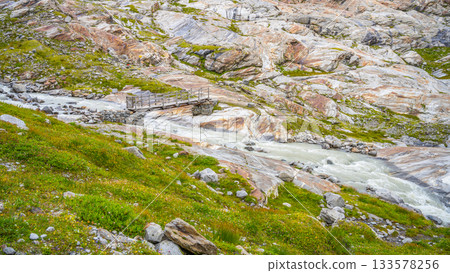 A wooden bridge spans a rushing stream sourced from the Schlaten Glacier, surrounded by rocky terrain and vibrant green vegetation in Hohe Tauern National Park. A wooden bridge spans a rushing stream sourced from the Schlaten Glacier, surrounded by rocky terrain and vibrant green vegetation in Hohe Tauern National Park. 133578256
