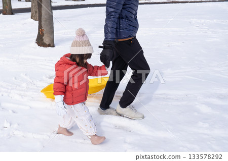 父親和孩子手牽手散步,孩子在雪橇上玩耍。 父親和孩子手牽手散步,孩子在雪橇上玩耍。 133578292