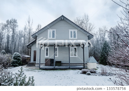 Wooden country house surrounded by snowy yard and trees in cold winter landscape. Calm rural scene with frost, snow, and seasonal atmosphere suitable for real estate, architecture, and winter themes. Wooden country house surrounded by snowy yard and trees in cold winter landscape. Calm rural scene with frost, snow, and seasonal atmosphere suitable for real estate, architecture, and winter themes. 133578756