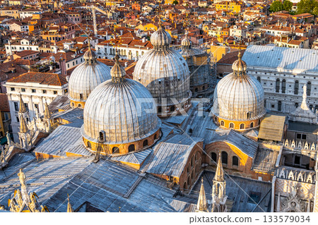 The ornate domes of St Marks Basilica tower above the picturesque rooftops of Venice, capturing the warm glow of sunset over the historic Piazza San Marco Square. The ornate domes of St Marks Basilica tower above the picturesque rooftops of Venice, capturing the warm glow of sunset over the historic Piazza San Marco Square. 133579034