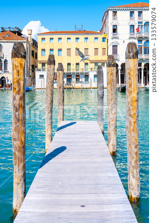A small wooden jetty extends into the calm turquoise waters of Venice, surrounded by charming buildings and the iconic atmosphere of this historic city under a clear blue sky. A small wooden jetty extends into the calm turquoise waters of Venice, surrounded by charming buildings and the iconic atmosphere of this historic city under a clear blue sky. 133579074
