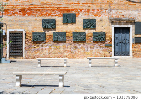 Bronze plaques commemorate Jewish history in the Venetian Ghetto, surrounded by empty benches against a vibrant brick wall in Venice, Italy. Bronze plaques commemorate Jewish history in the Venetian Ghetto, surrounded by empty benches against a vibrant brick wall in Venice, Italy. 133579096
