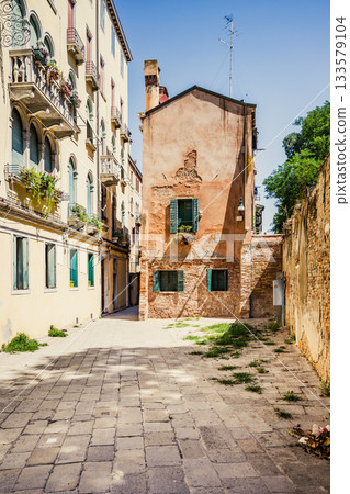 A peaceful alleyway in Venice showcases beautiful architecture with a weathered building and vibrant greens. The sun illuminates the street, highlighting its old-world charm and quaint atmosphere. A peaceful alleyway in Venice showcases beautiful architecture with a weathered building and vibrant greens. The sun illuminates the street, highlighting its old-world charm and quaint atmosphere. 133579104