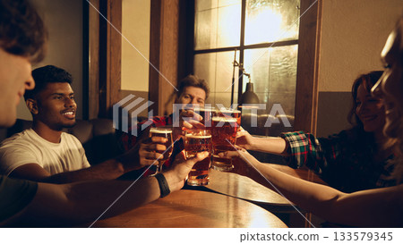 Friends raising beer glasses together at pub booth in warm scene 133579345