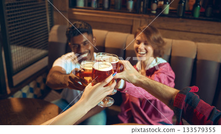 Friends clinking beer glasses with joyful expressions in pub Friends clinking beer glasses with joyful expressions in pub 133579355