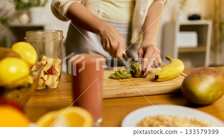 Hands slicing ripe kiwi for smoothie preparation with fruits and blender on table 133579399