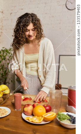 Young woman preparing fruit for smoothie with fresh produce and blender on wooden table 133579403