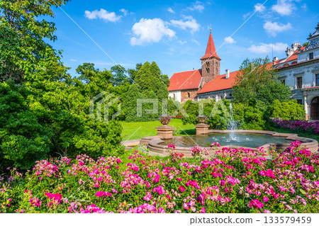 Surrounded by vibrant flower beds and lush greenery, the Church of the Nativity of the Virgin Mary stands tall in Pruhonice Castle, showcasing its stunning architecture against a bright blue sky. Surrounded by vibrant flower beds and lush greenery, the Church of the Nativity of the Virgin Mary stands tall in Pruhonice Castle, showcasing its stunning architecture against a bright blue sky. 133579459