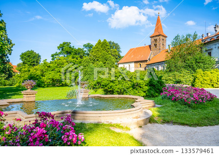 The Church of the Nativity of the Virgin Mary in Pruhonice Castle stands amid lush greenery and vibrant flowers. A tranquil fountain enhances the serene atmosphere on a sunny day. 133579461