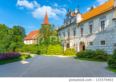 Pruhonice Castle showcases the stunning Church of the Nativity of the Virgin Mary, surrounded by vibrant greenery and colorful flowers under a clear blue sky. Visitors admire the architecture. Pruhonice Castle showcases the stunning Church of the Nativity of the Virgin Mary, surrounded by vibrant greenery and colorful flowers under a clear blue sky. Visitors admire the architecture. 133579463