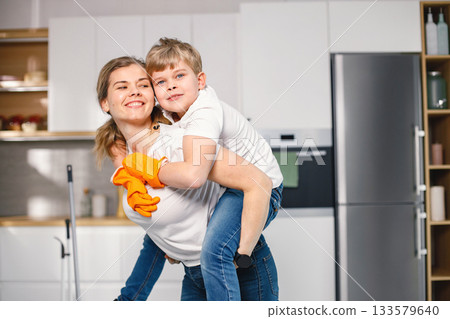 Blonde boy and adult woman cleaning in a kitchen and wearing orange gloves. Caucasian boy helping his mother to do housework. Mother carrying her son piggyback. Blonde boy and adult woman cleaning in a kitchen and wearing orange gloves. Caucasian boy helping his mother to do housework. Mother carrying her son piggyback. 133579640
