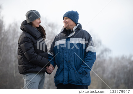 Two friends man and woman warmly dressed, holding hands while walking along snowy path in winter landscape. Cold season. Outdoor. Friendship, love and warmth on cold day. Two friends man and woman warmly dressed, holding hands while walking along snowy path in winter landscape. Cold season. Outdoor. Friendship, love and warmth on cold day. 133579947