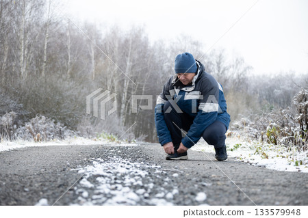 Adult sporty man at walk in winter clothing tying shoelaces and snowy road at park. Cold weather and snowy season. Outdoors. Adult sporty man at walk in winter clothing tying shoelaces and snowy road at park. Cold weather and snowy season. Outdoors. 133579948