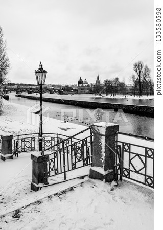 Winter scene on the Vltava riverbank in Prague. It features a staircase leading down to the water, adorned with a decorated railing and a historical lamp. In the background, there is a view of the Winter scene on the Vltava riverbank in Prague. It features a staircase leading down to the water, adorned with a decorated railing and a historical lamp. In the background, there is a view of the 133580598