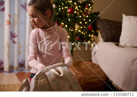 Girl Packing Backpack Near Christmas Tree in Cozy Living Room During Holidays 133580711