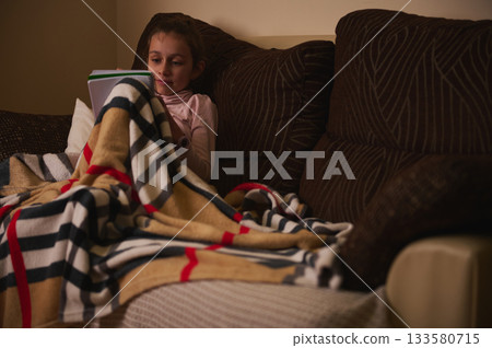 Girl Reading Tablet Under Cozy Blanket on Couch, Family Night at Home 133580715