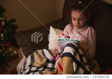 Child Writing in Notebook on Couch With Blanket Near Christmas Tree Lights Child Writing in Notebook on Couch With Blanket Near Christmas Tree Lights 133580716