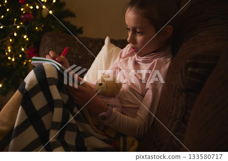 Child Reading Hobby By Christmas Tree On Sofa With Teddy Bear And Cozy Blanket Child Reading Hobby By Christmas Tree On Sofa With Teddy Bear And Cozy Blanket 133580717