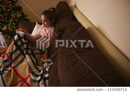 Cozy Christmas Night: Girl Reading on Couch with Blanket and Teddy Bear Near Tree Cozy Christmas Night: Girl Reading on Couch with Blanket and Teddy Bear Near Tree 133580718