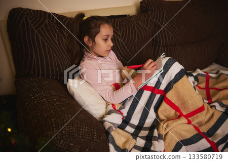Cozy Girl Writes in Notebook While Snug Under Blanket on Couch 133580719