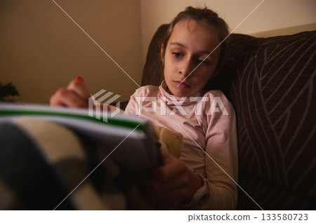 Girl Reading Book on Couch at Night Creates Cozy Home Moment Girl Reading Book on Couch at Night Creates Cozy Home Moment 133580723
