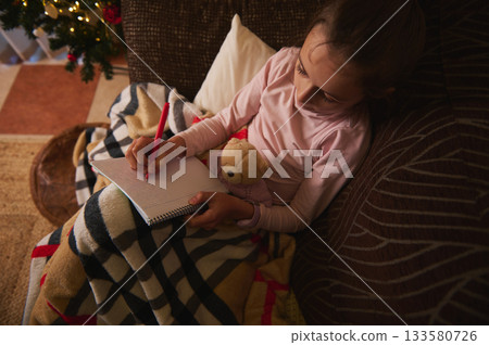 Little Girl Writing in Notebook on Sofa with Teddy Bear and Cozy Blanket by Christmas Tree 133580726