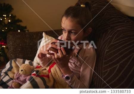 Young Girl Relaxing On Couch With Wooden Toy Instrument Near Christmas Tree Young Girl Relaxing On Couch With Wooden Toy Instrument Near Christmas Tree 133580733