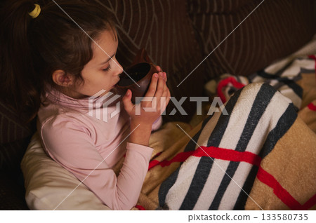 Young Girl Sips Drink From Cup While Resting On Couch With Blanket Young Girl Sips Drink From Cup While Resting On Couch With Blanket 133580735