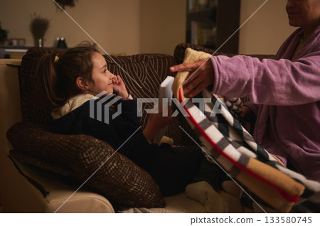Young Girl Reading a Book on Sofa as Grandparent Covers Her with a Blanket in Cozy Living Room 133580745